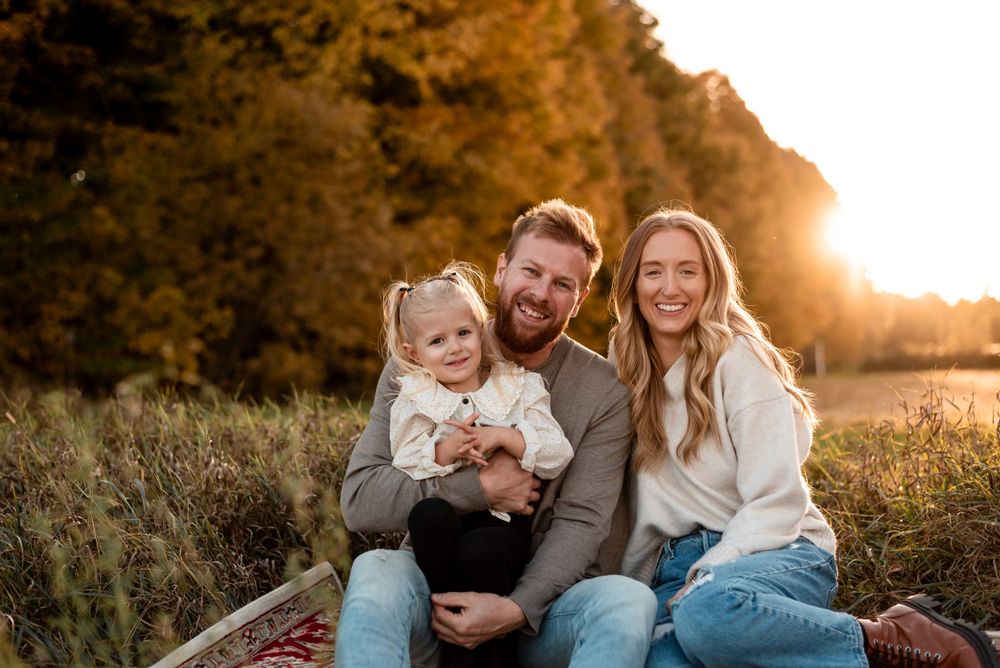 Family sits together on a blanket outdoors during golden hour sunset with autumn trees in background.
