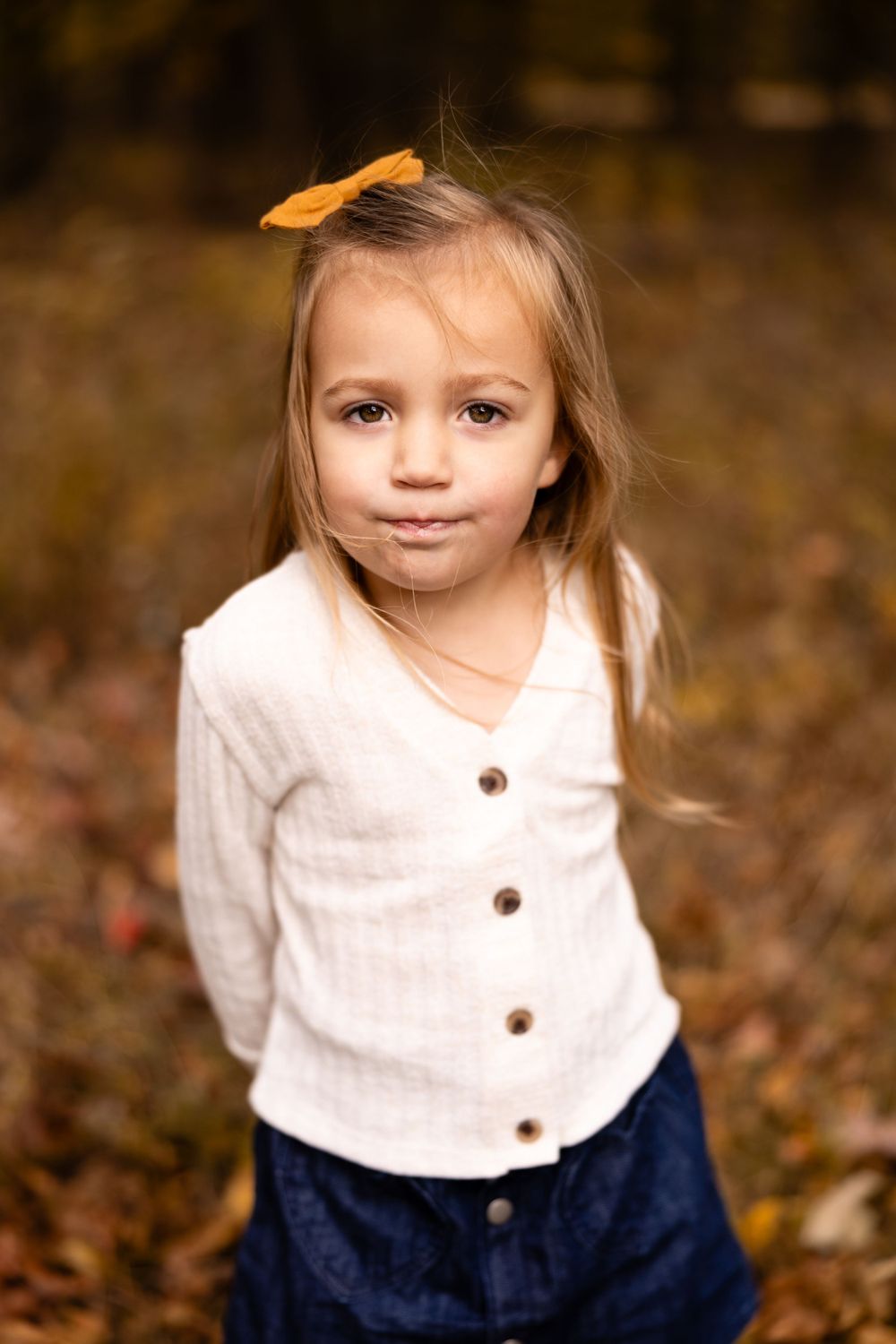 Adorable little girl with orange bow in autumn forest wearing white button-up top.