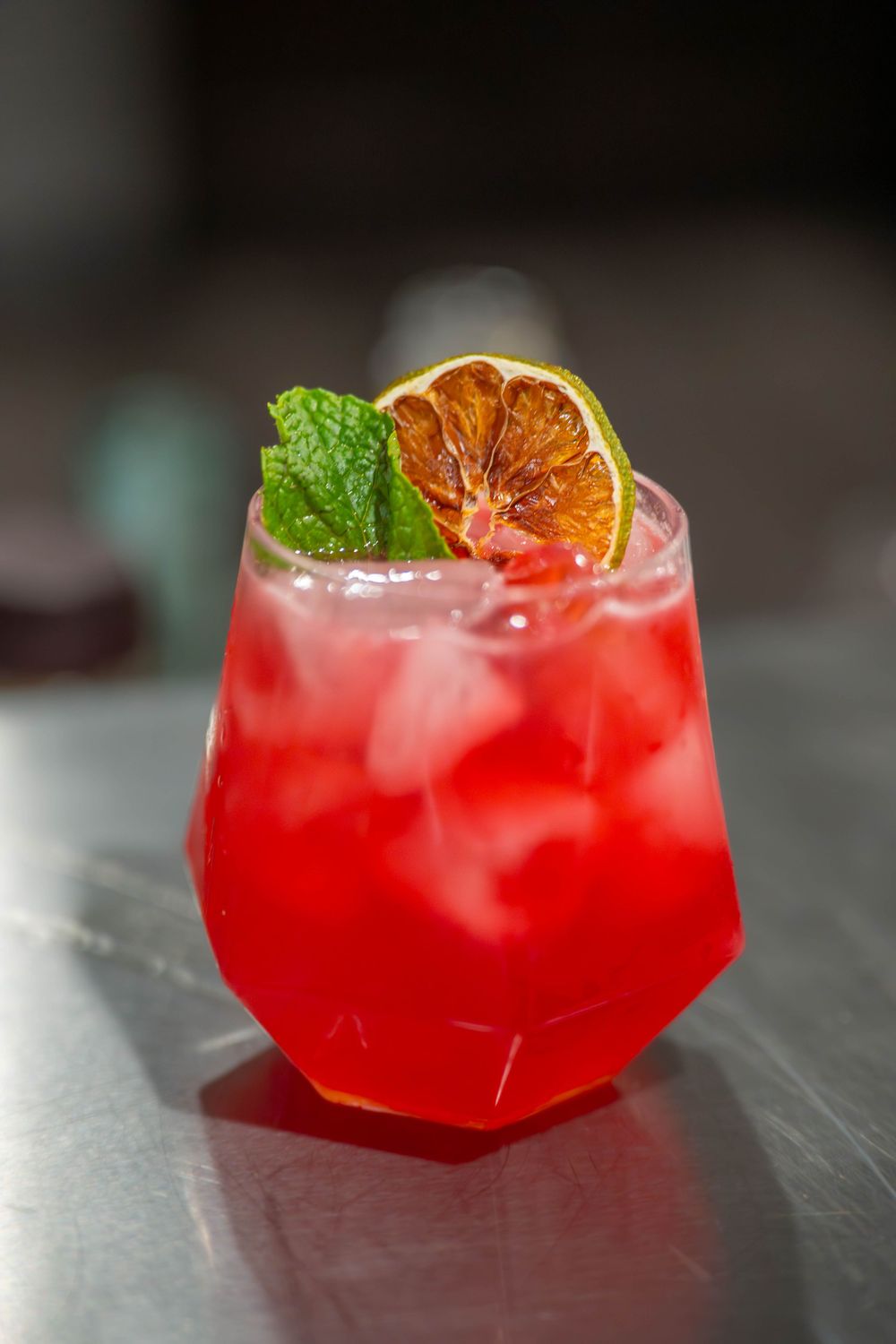 Red cocktail with ice, dried lime slice, and fresh mint garnish in a geometric glass on a bar surface. Quad Cities Product Photography