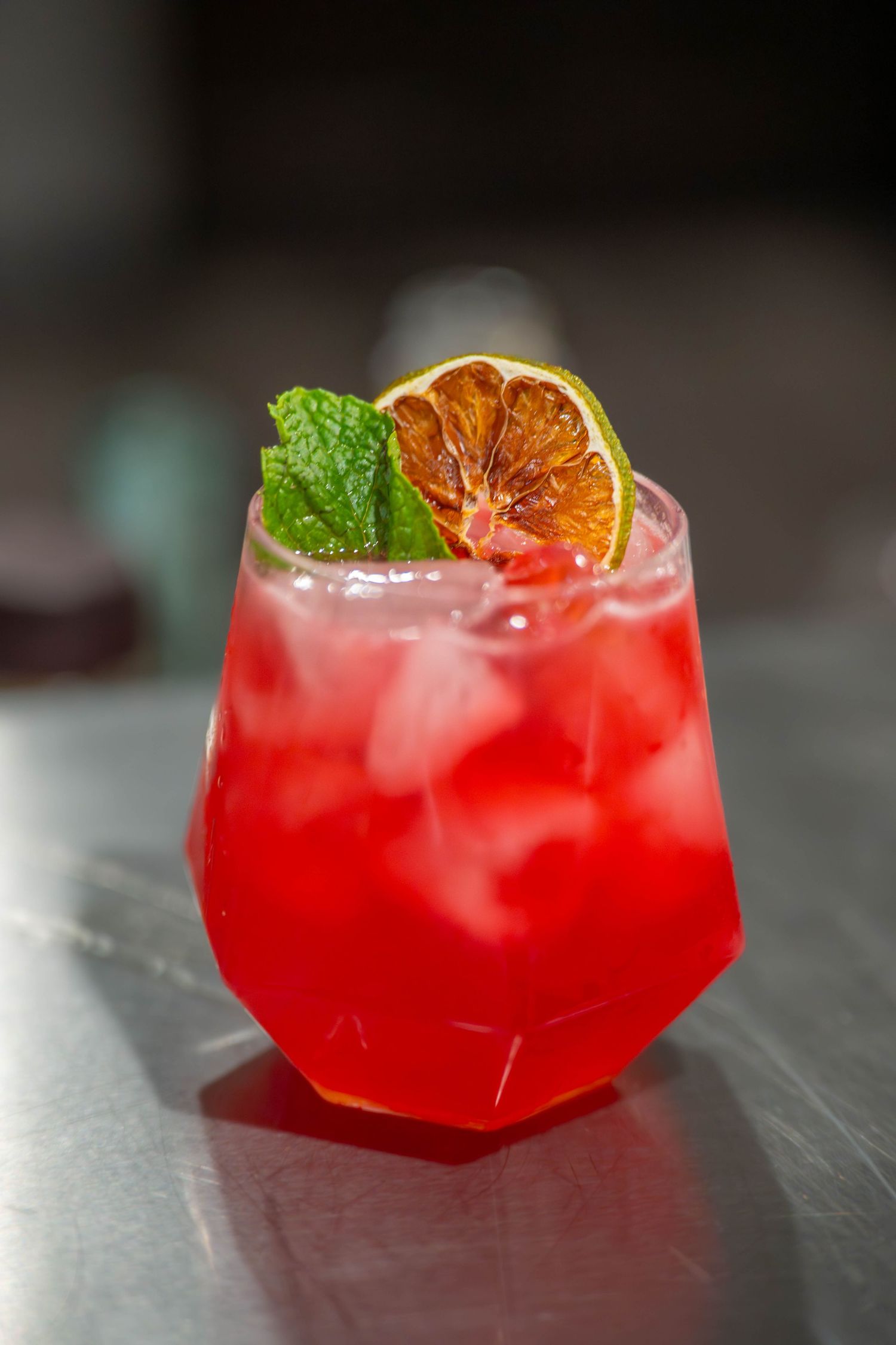 Red cocktail with ice, dried lime slice, and fresh mint garnish in a geometric glass on a bar surface. Quad Cities Product Photography