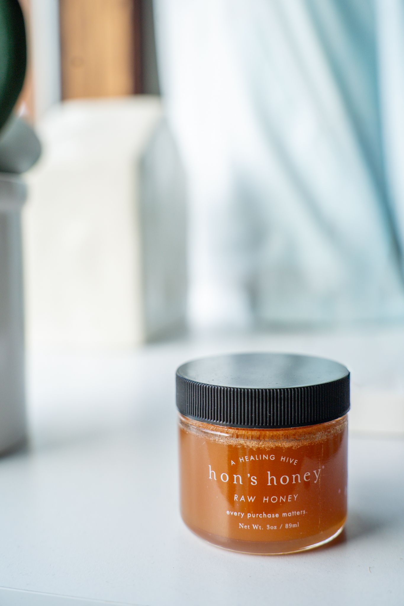 A jar of raw Hon's honey with black lid sits on white surface with soft blurred background. Quad Cities Product Photography Argrow's House