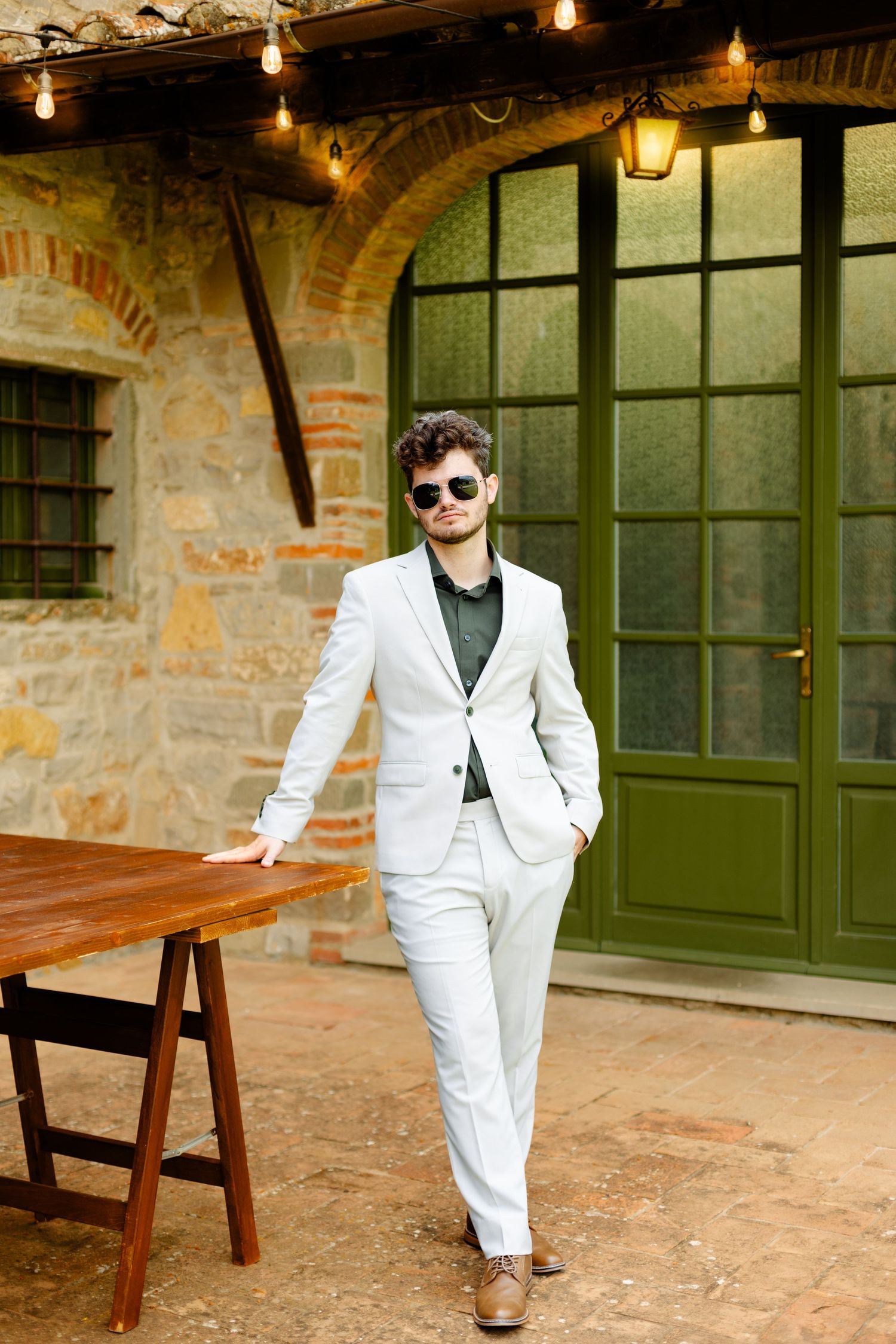 A person in a white suit poses confidently against a rustic stone building with string lights.