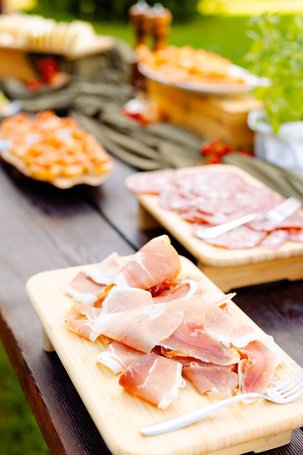 Charcuterie board with sliced ham and meats arranged on wooden serving plank outdoors.