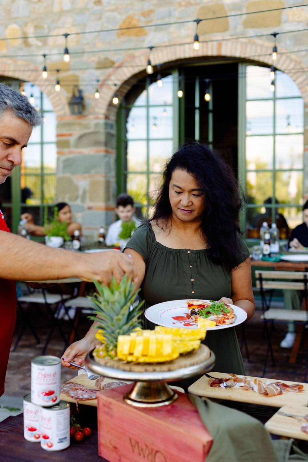 Two people serve fresh fruit and dishes at an outdoor restaurant patio with stone walls and string lights.
