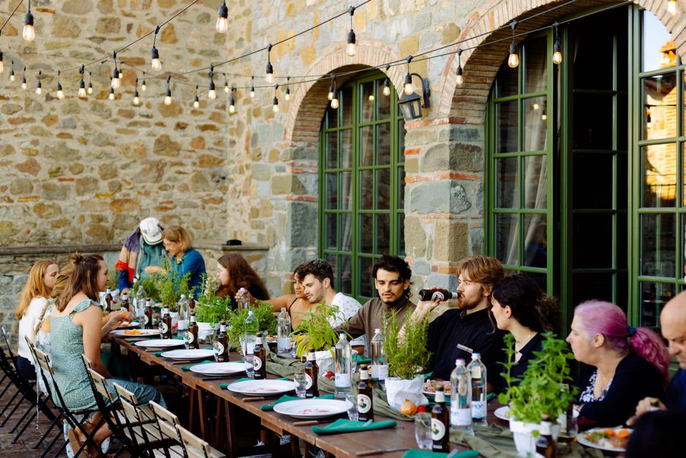 A group dines al fresco at a long table under string lights against a rustic stone building.