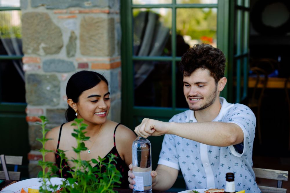 Two diners share a conversation at an outdoor restaurant table with stone walls.