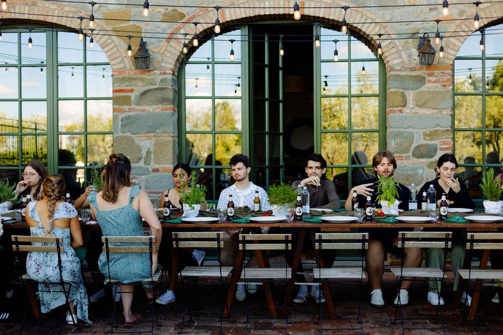Diners gather at a long communal table on an outdoor patio with stone walls and arched windows.