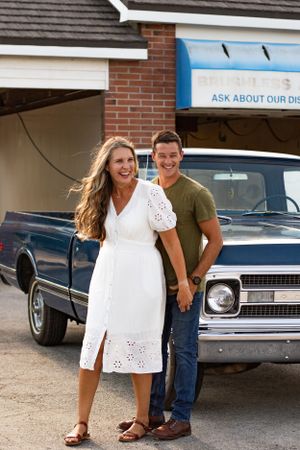 Couple standing next to classic cars outside a brick building with a white dress and casual attire.