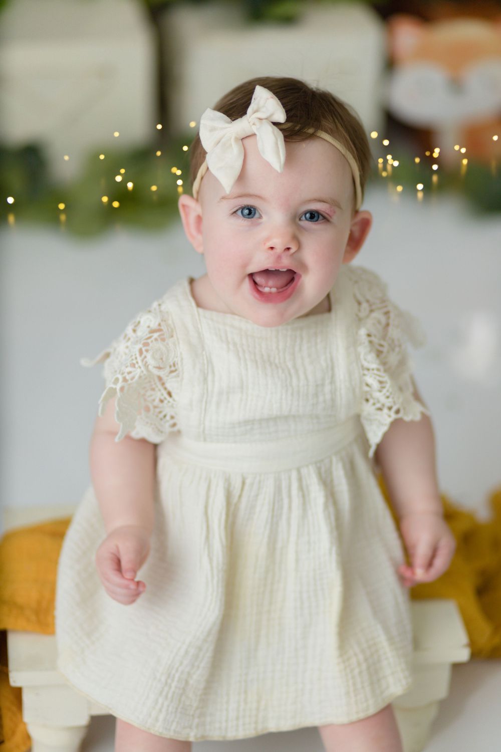 A small child in a cream-colored dress with lace details and white bow smiles at camera against a sparkly backdrop.