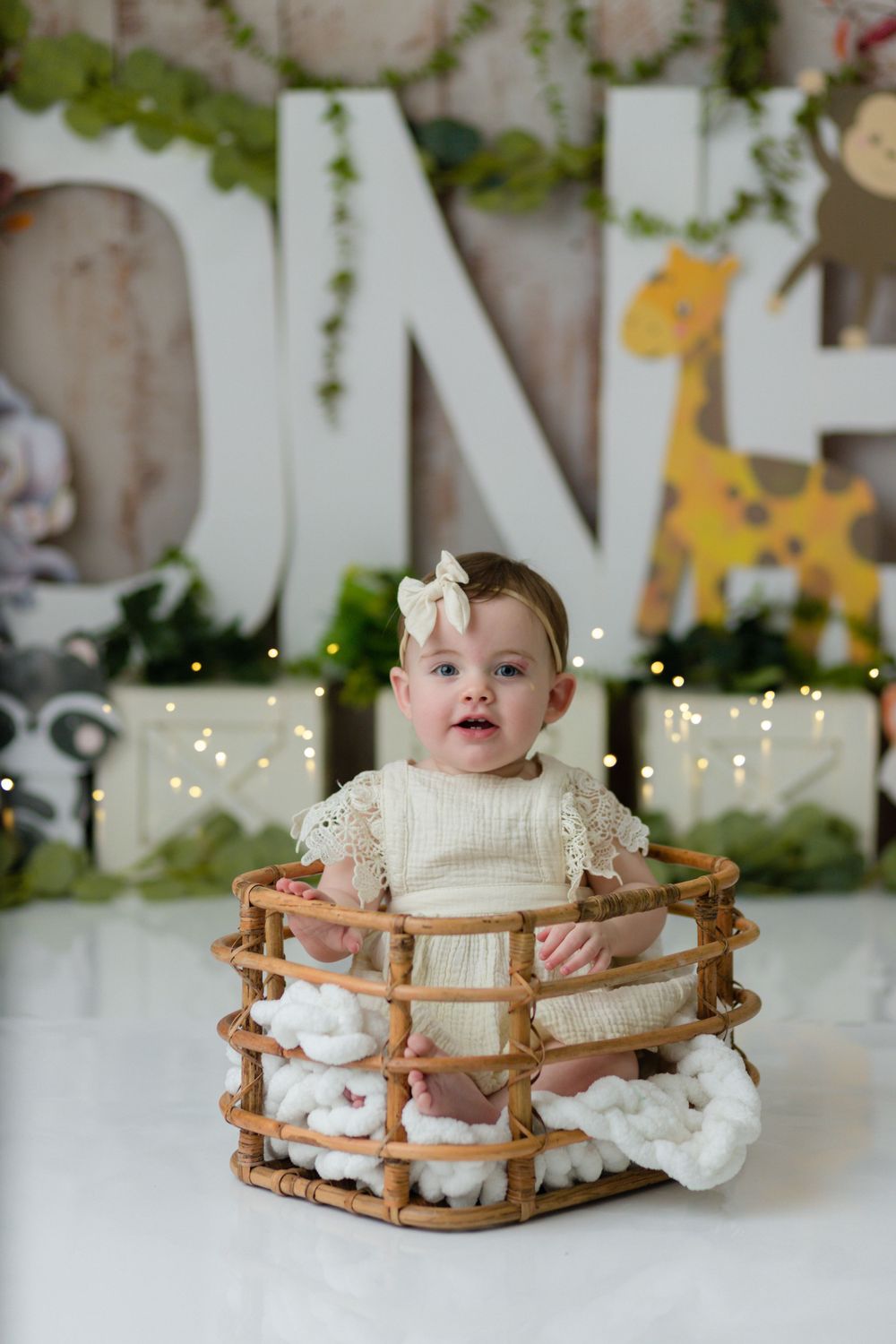A small child in cream dress sits inside a wooden basket prop against a decorated background.