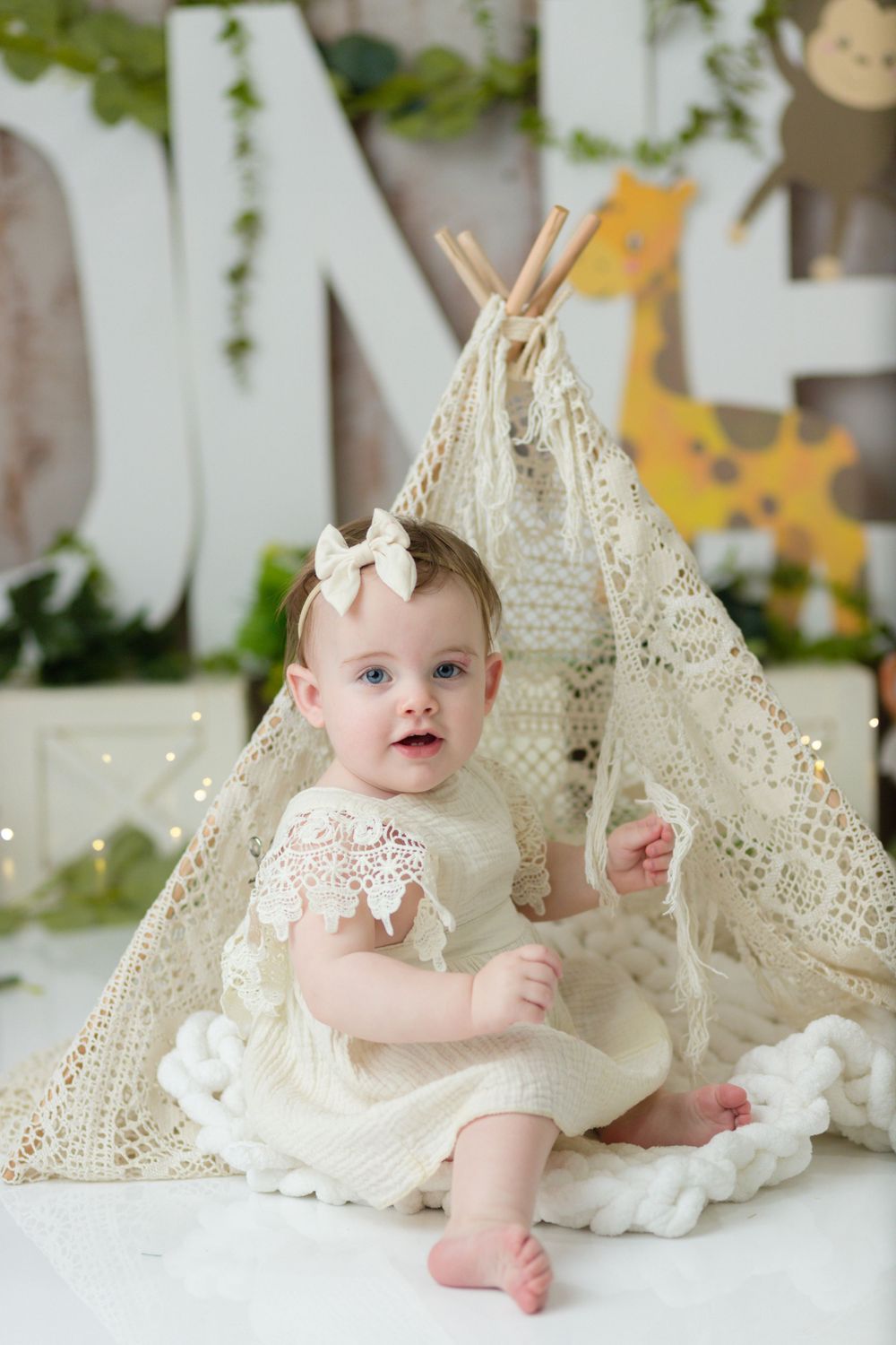 A small child in lace dress sits by a decorative teepee with fairy lights and woodland backdrop.