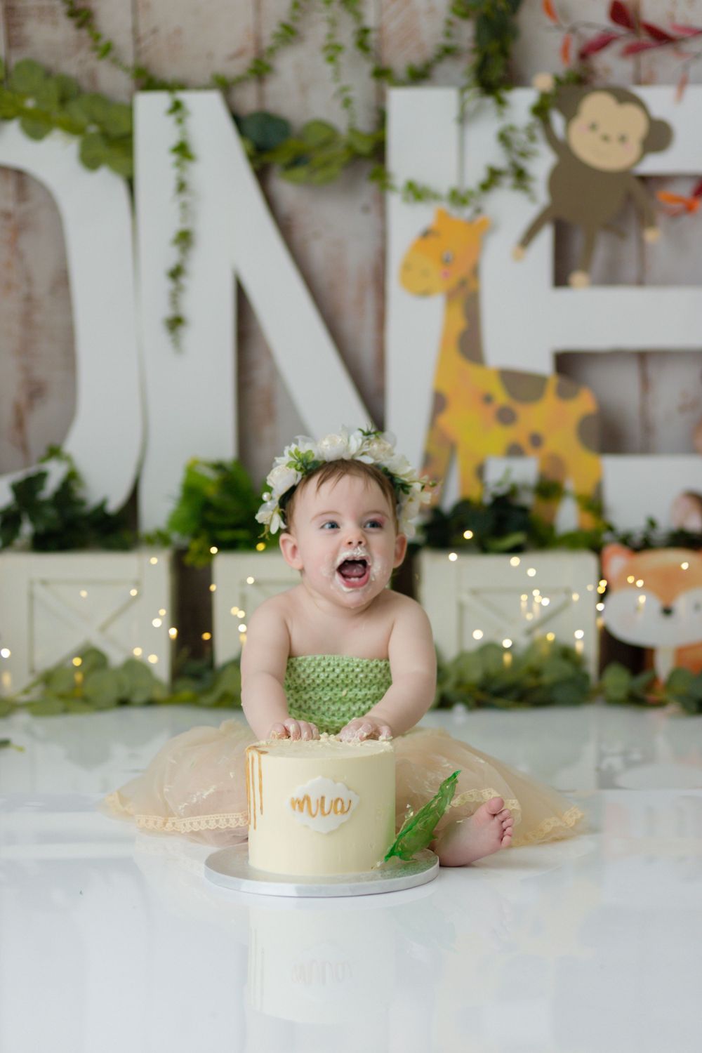 A delighted toddler in green tulle dress sits with a small cake during a photo shoot.