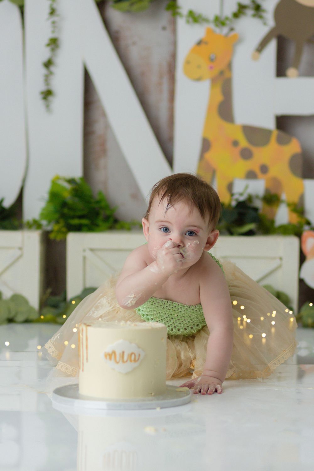 A messy-faced toddler in green outfit reaches for a small cream-colored cake.