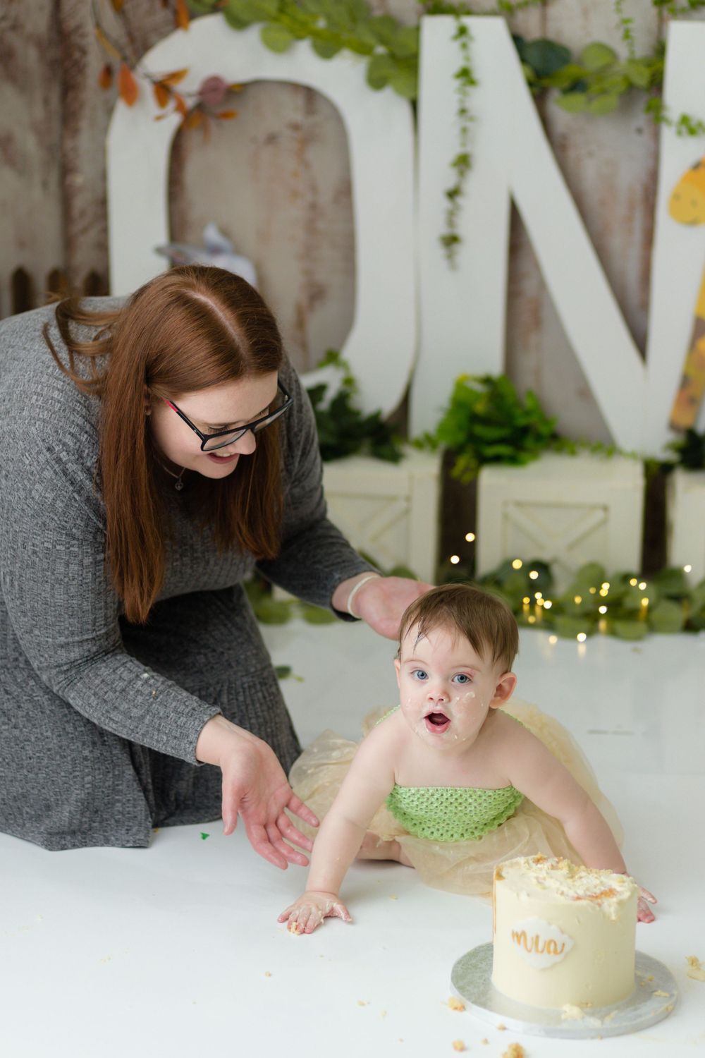 A cake smash photo session with a small child and caregiver against a decorated backdrop.