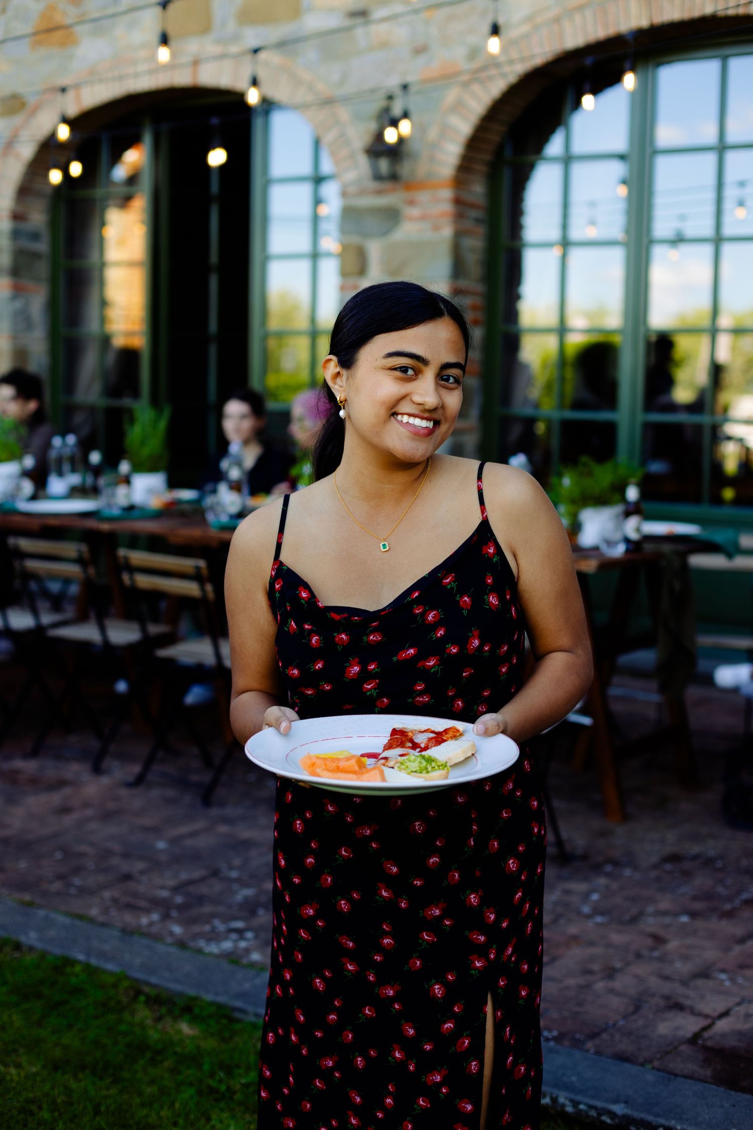 Person in black dress with red pattern holding plate of food at outdoor dining venue.