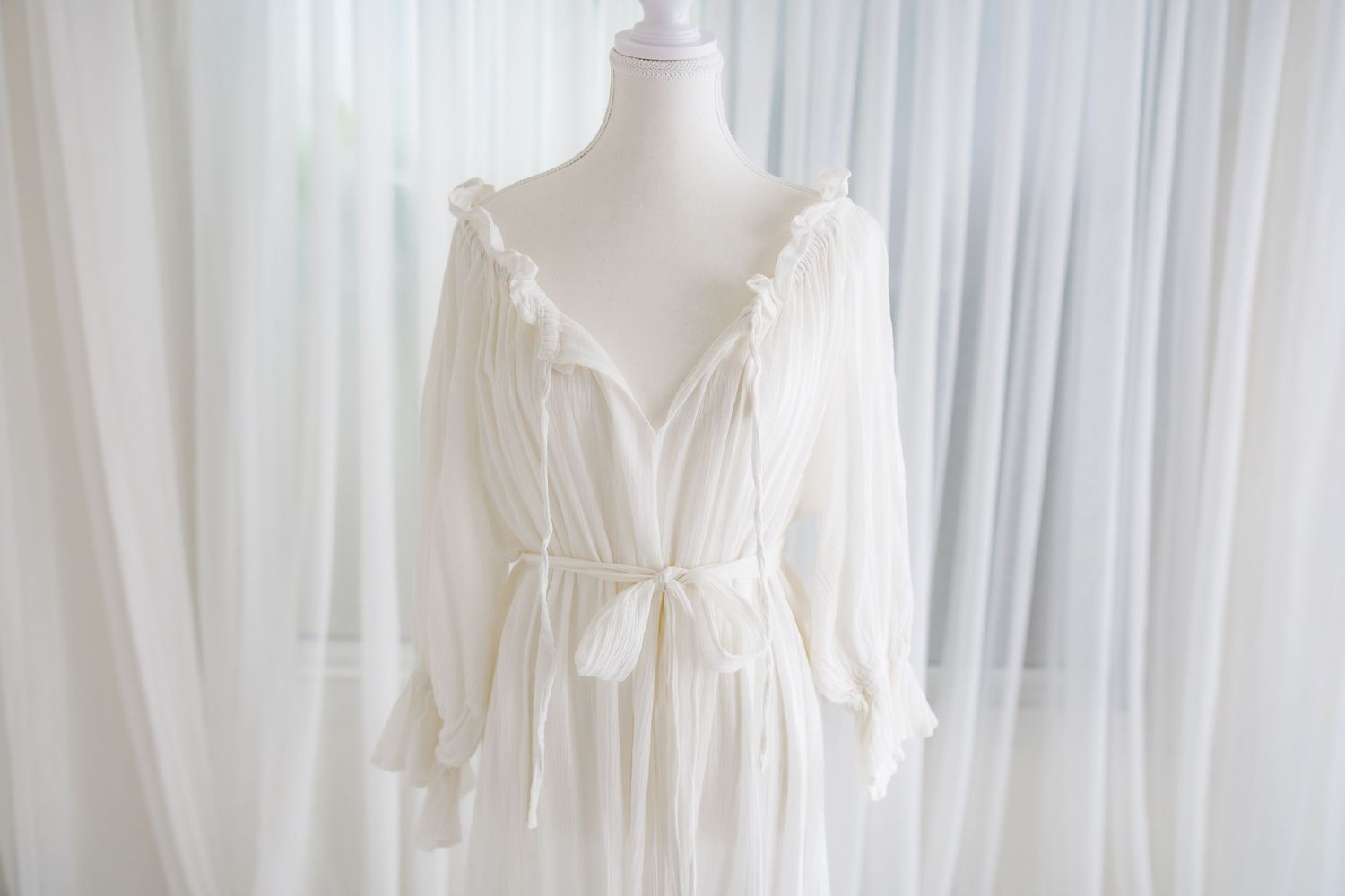 Romantic white cotton dress with V-neckline and flowing sleeves displayed against curtains.