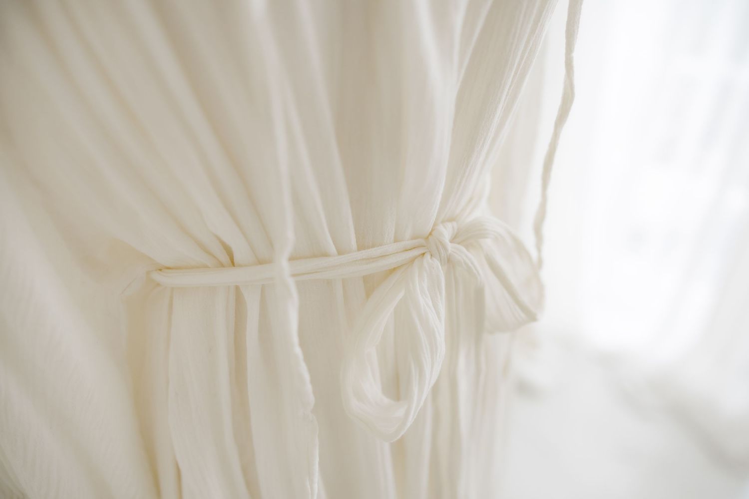 Detail shot of white cotton dress tie waist against sheer curtain background.