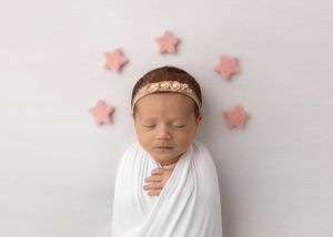 Peaceful newborn in white swaddle with pink stars and delicate headband on white backdrop.