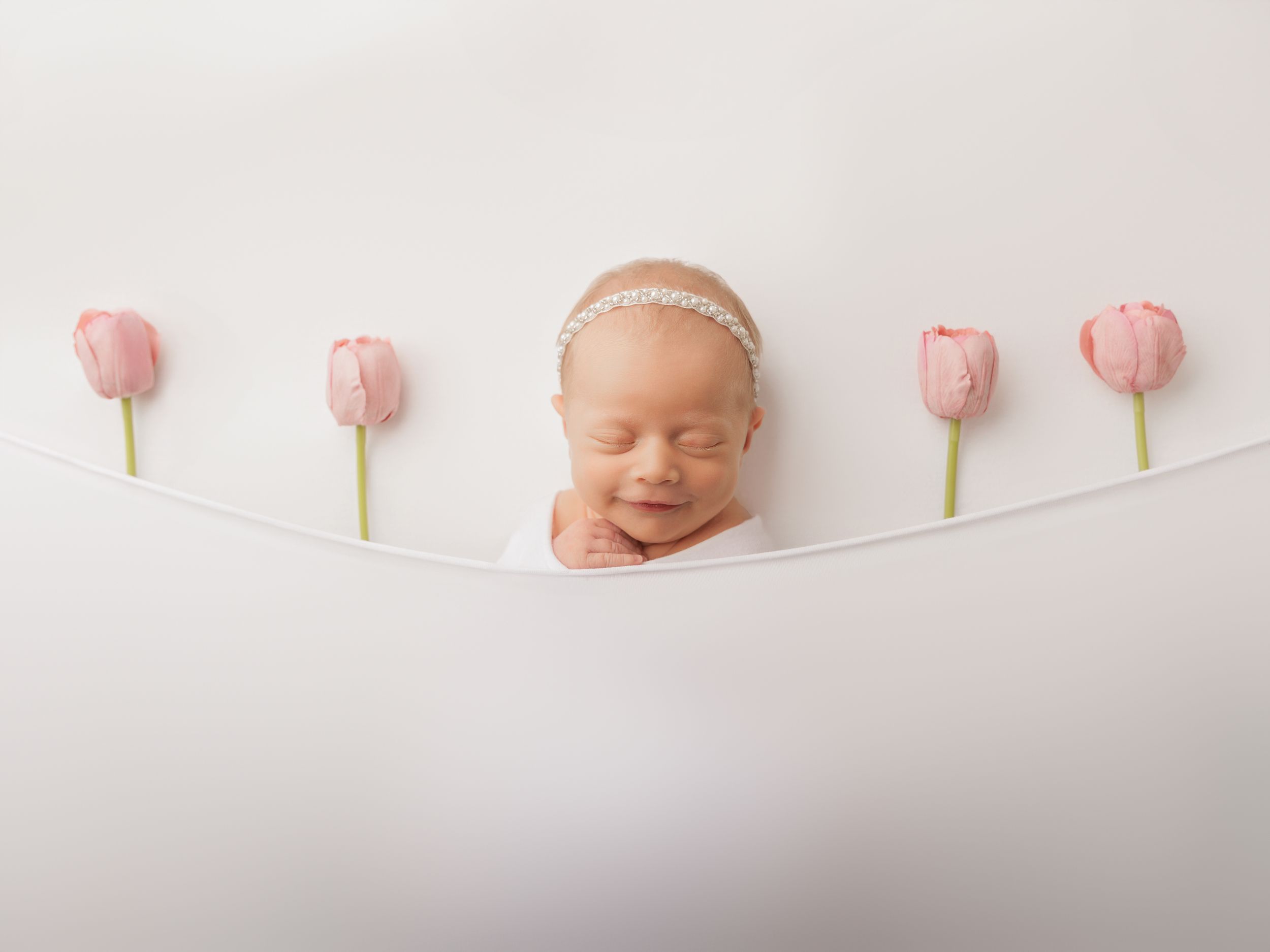 A sleeping newborn with closed eyes is nestled between four pink tulips on a white backdrop in a minimalist composition.