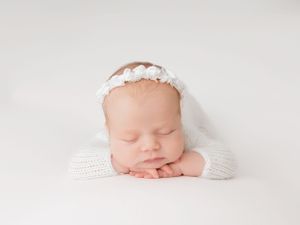 Sleeping newborn wearing white lace headband rests peacefully on white blanket in soft lighting.