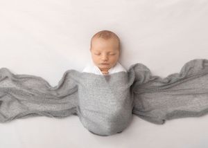 Sleeping newborn swaddled in soft gray wrap with flowing fabric wings against white background.