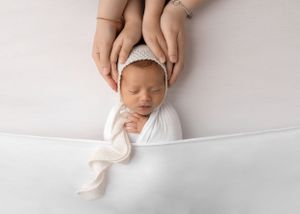 Sleeping newborn in white wrap with gentle hands cradling head against white background.