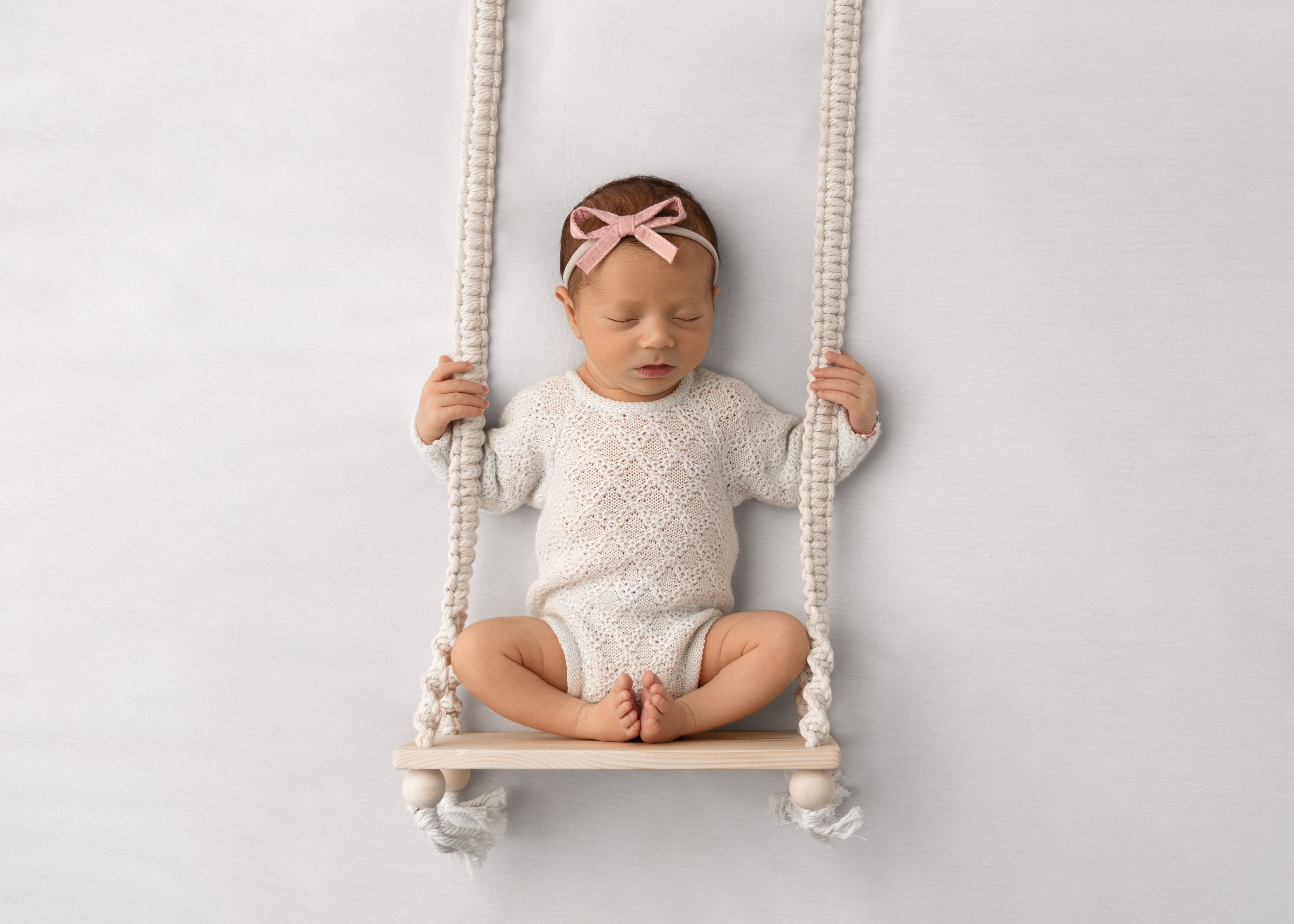 Newborn in white outfit sitting on rustic wooden swing with macrame ropes.
