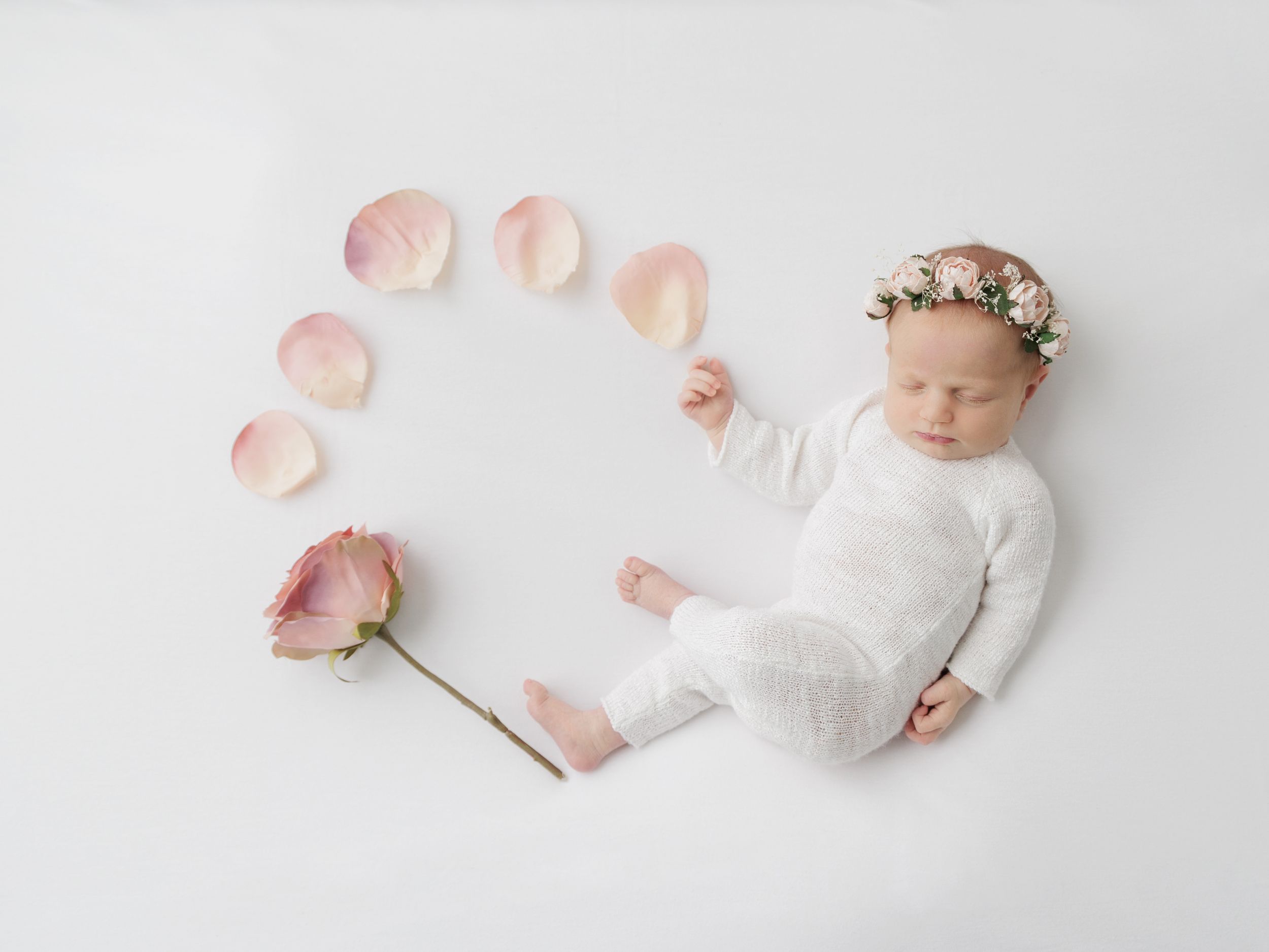 Sleeping newborn in white with floral crown surrounded by pink rose petals in artistic composition.