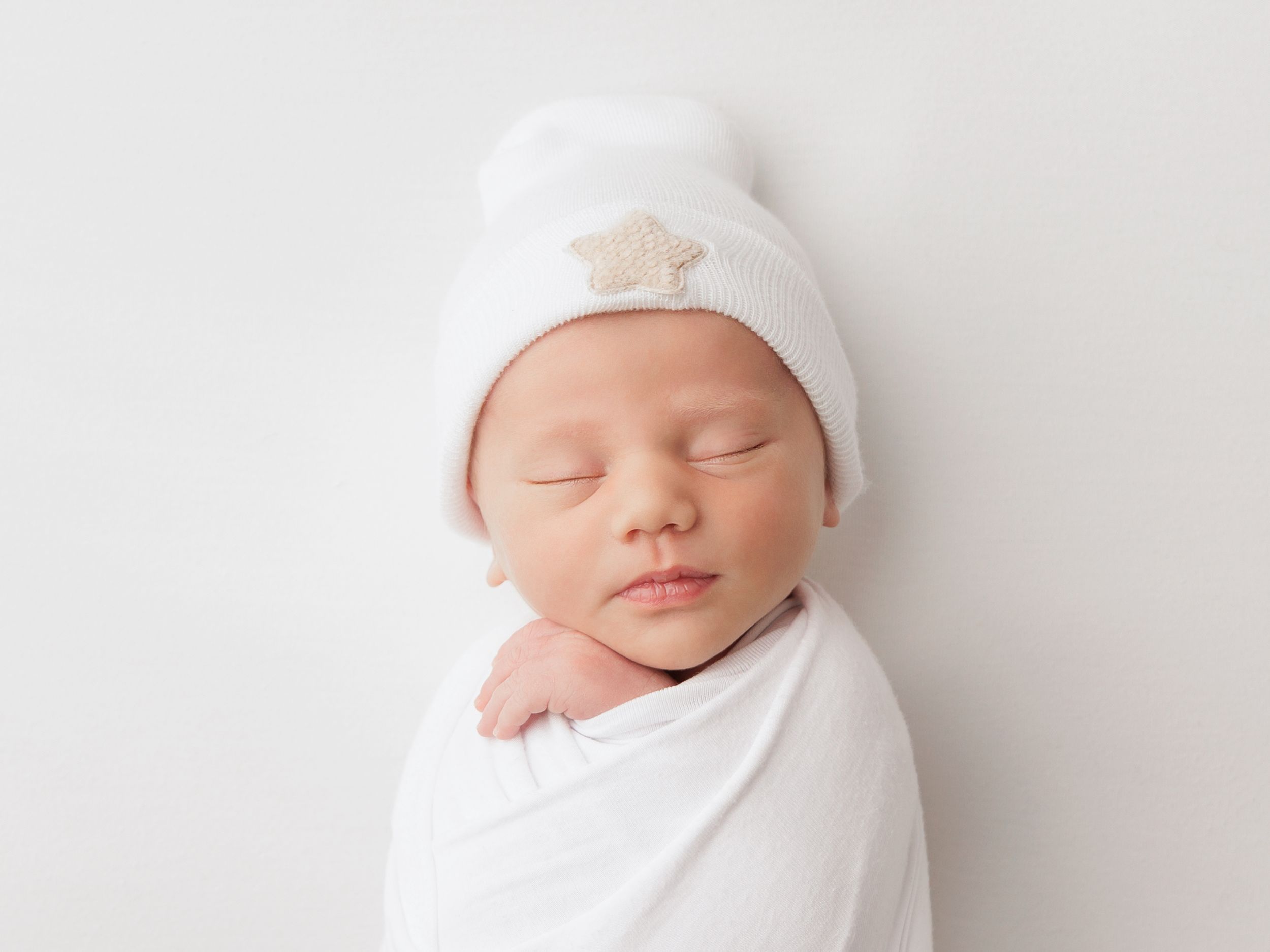 A sleeping newborn baby swaddled in white fabric with a matching white hat rests peacefully.
