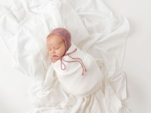 Sleeping newborn wrapped in white with pink knit bonnet on textured white fabric.