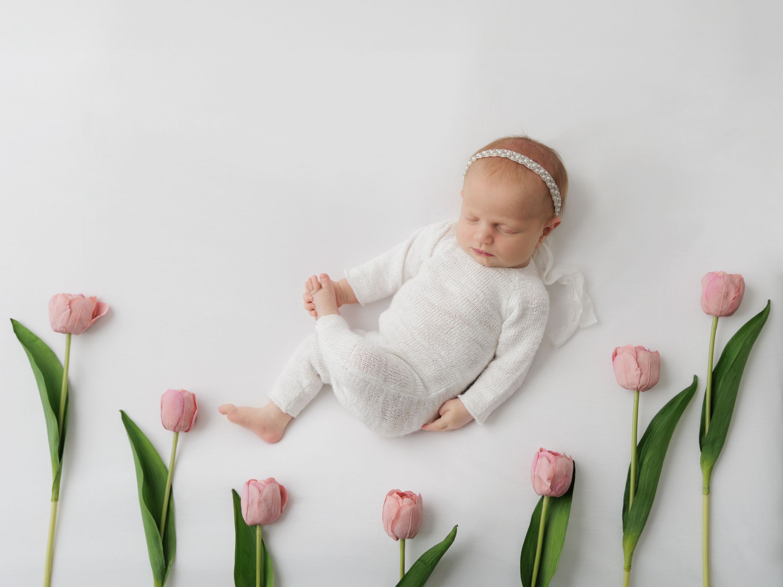 Sleeping baby in white outfit surrounded by pink tulips in minimalist photo composition.