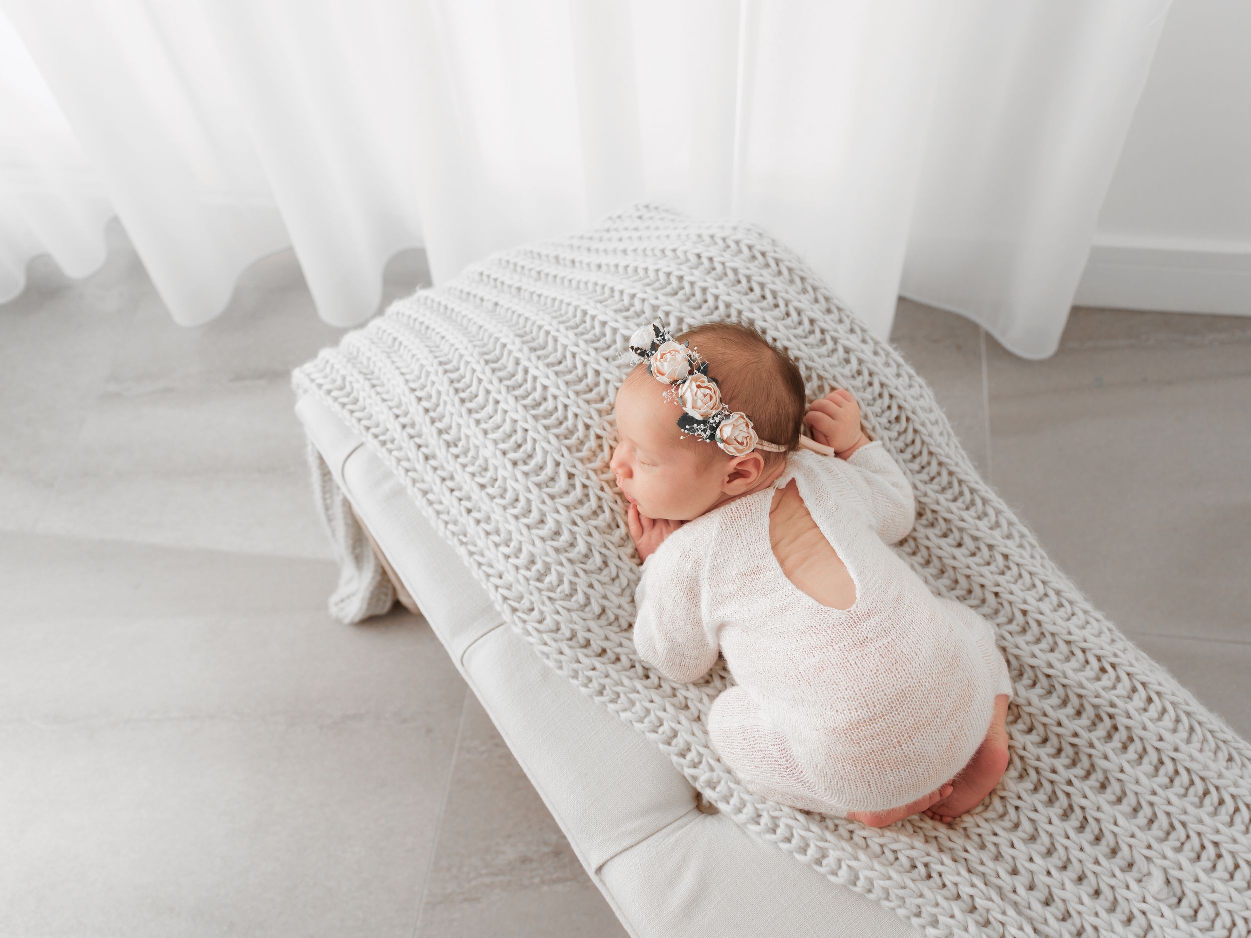Newborn baby in white onesie sleeping peacefully on gray knitted blanket in bright room.