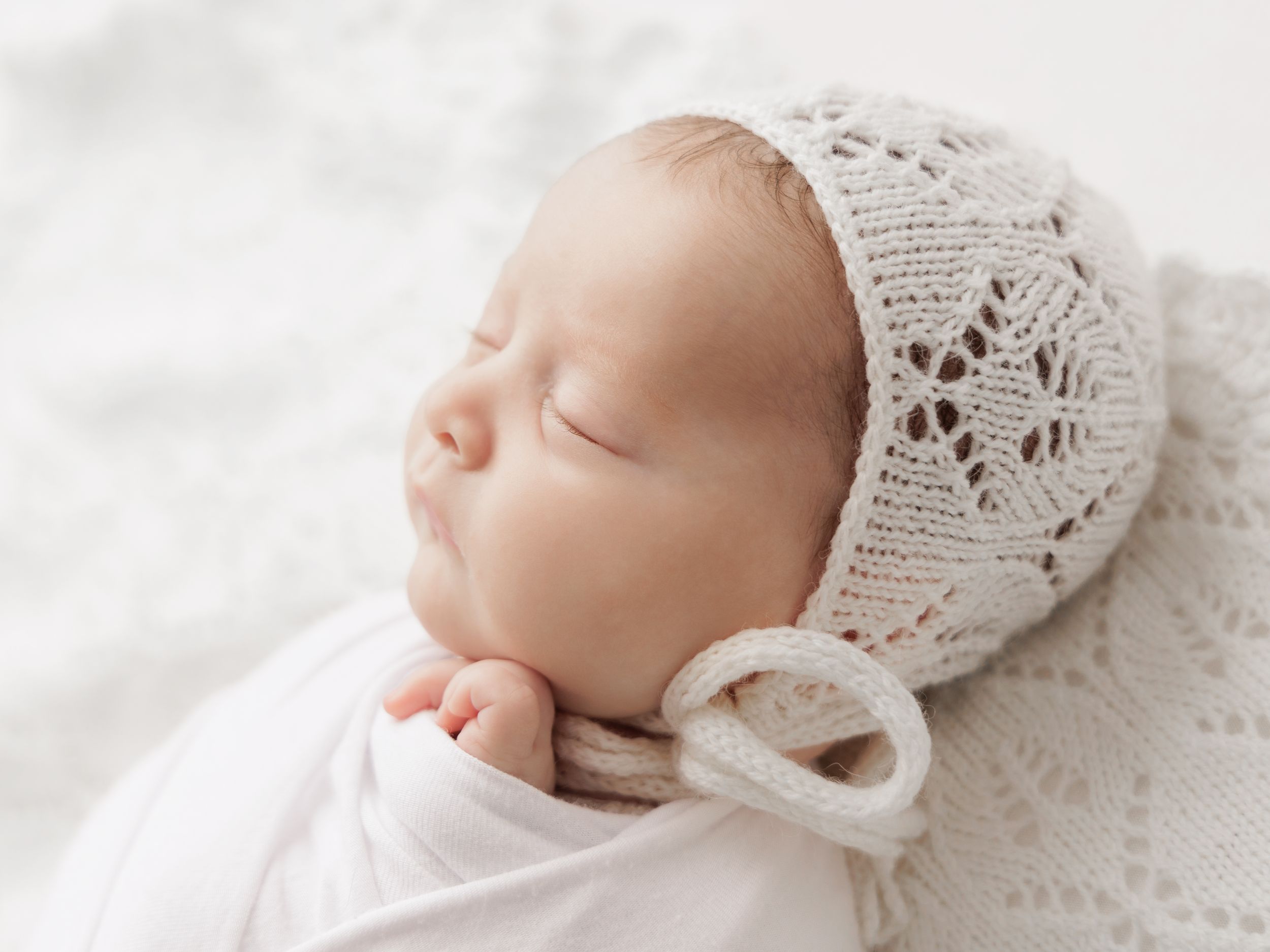 Sleeping newborn wearing a delicate white lace bonnet and swaddled in soft white fabric during professional photoshoot.