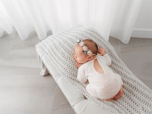 Newborn baby in white onesie sleeping peacefully on gray knitted blanket in bright room.