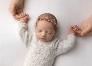 Sleeping newborn in white knit outfit with floral headband held by parents' hands.