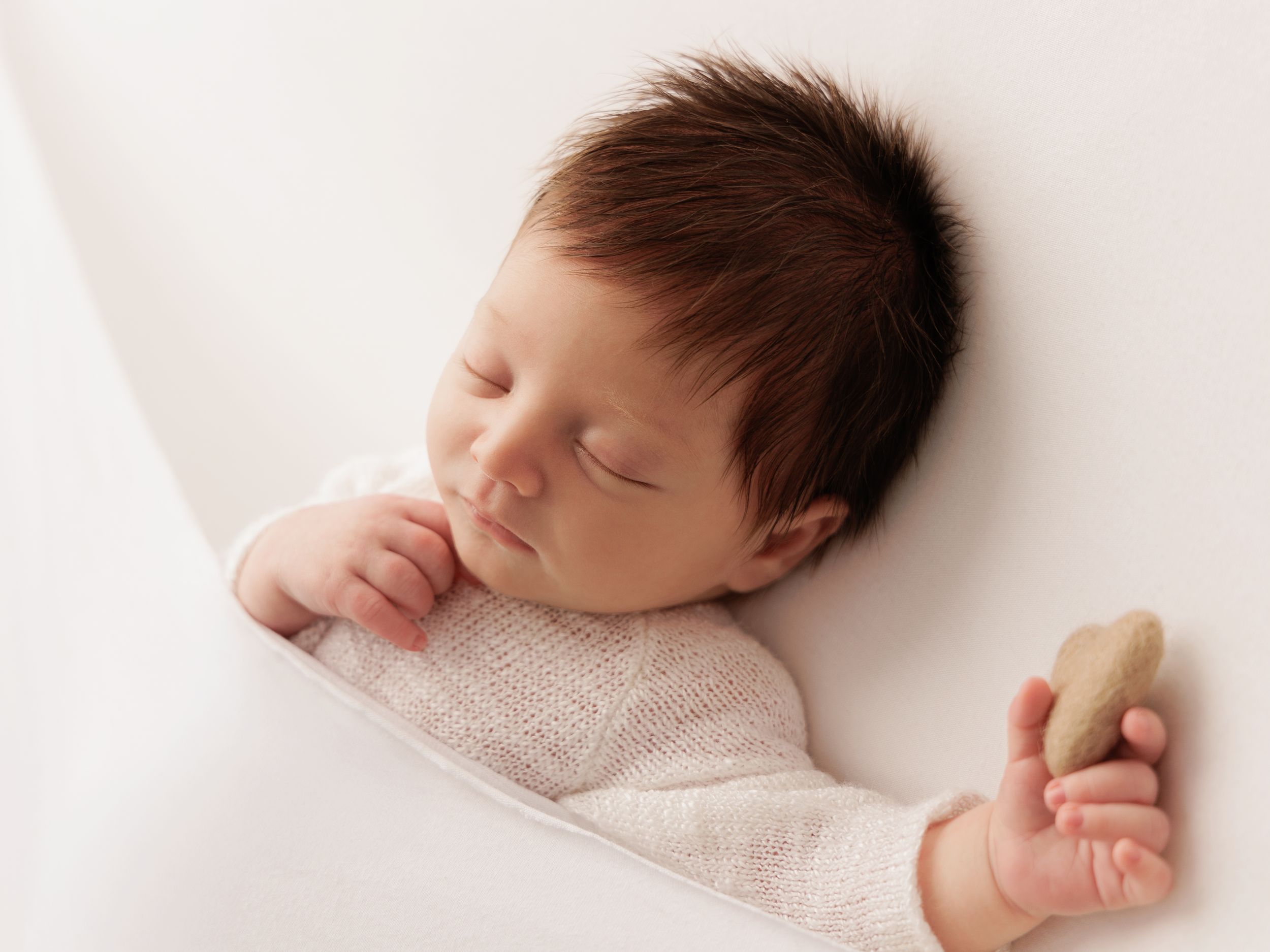Peaceful sleeping baby in white knit outfit holding a soft plush toy against pale background.