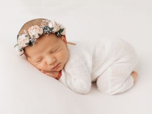 A sleeping newborn wearing a floral crown and white onesie on a white background.
