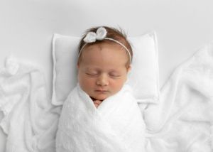 Sleeping newborn wrapped in white swaddle with bow headband on white pillow.