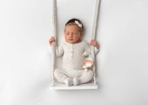 Baby in white knit outfit sitting on wooden swing with rope details.