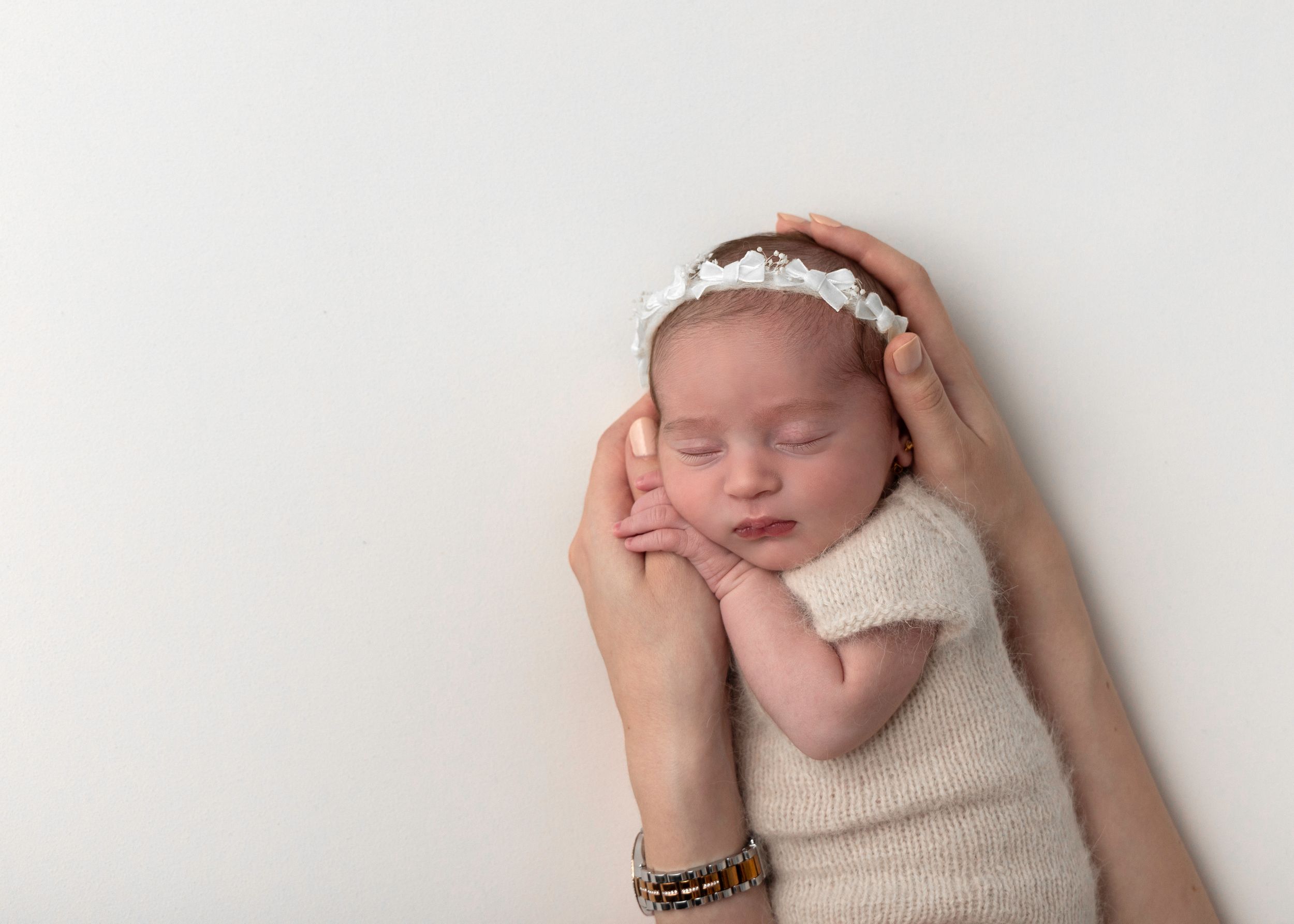 Sleeping newborn in cream wrap with floral headband being gently cradled.