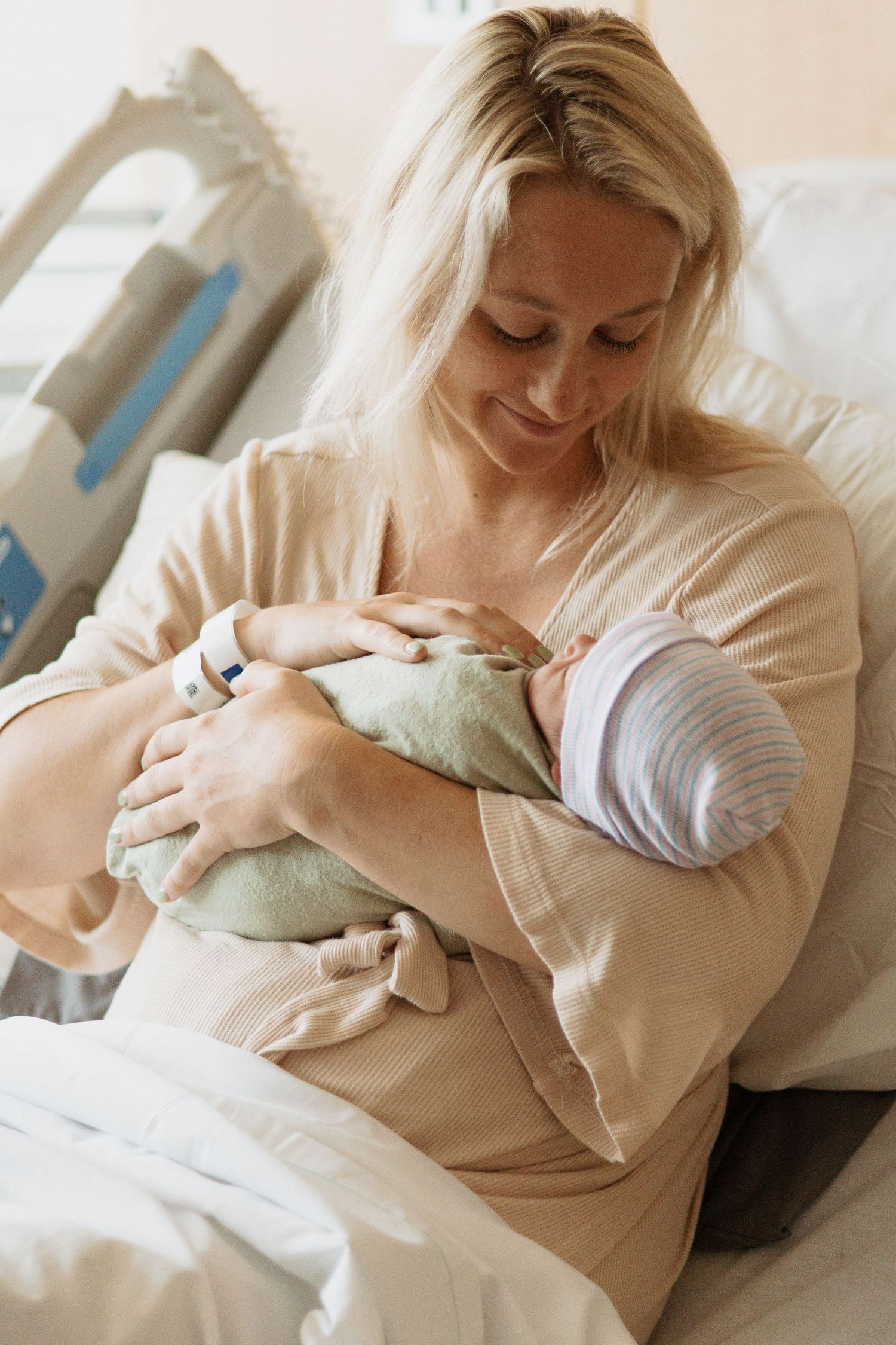 New parent in hospital bed cradles newborn wrapped in mint green swaddle.