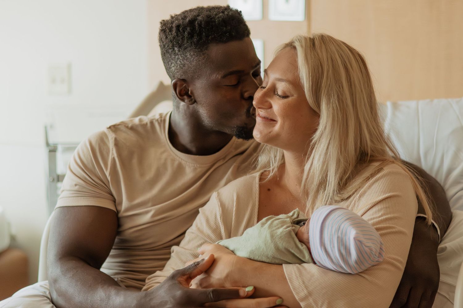 A couple in beige clothing shares an intimate moment together with their newborn baby on a couch.