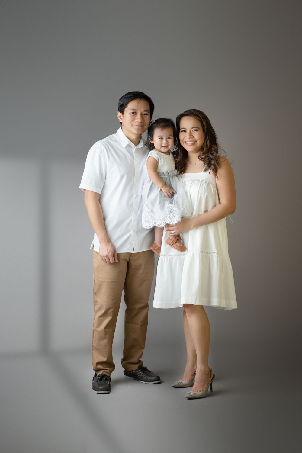 Studio family portrait with three people in coordinated white and beige outfits against grey backdrop.
