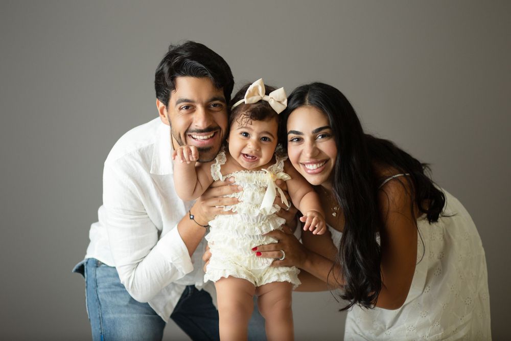 Family portrait of three people wearing white outfits smiling and laughing together against grey background.