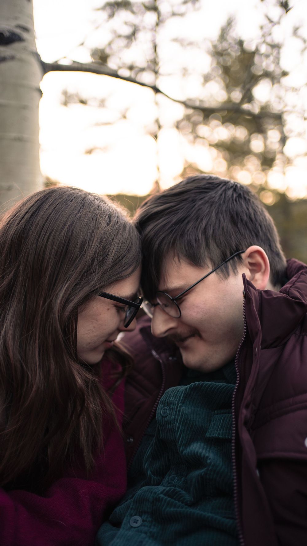 A tender moment shared between two people wearing glasses as they lean in close together outdoors at sunset.