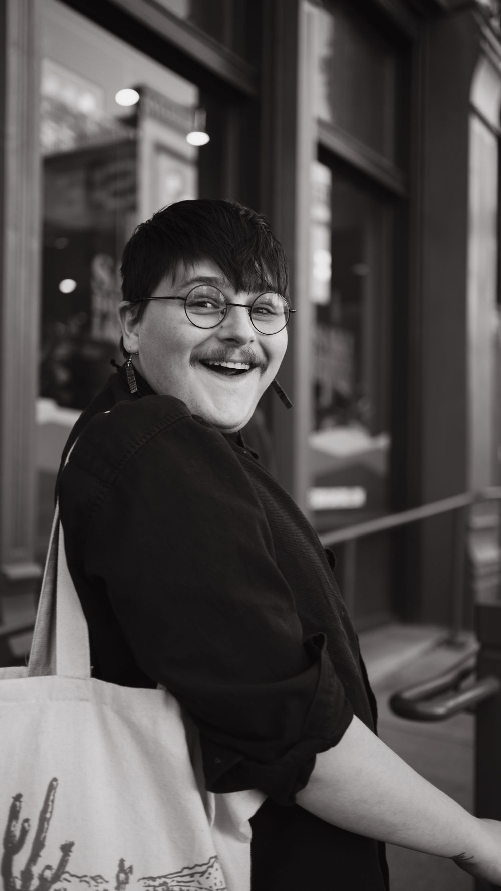 Person in black attire smiling while carrying a tote bag outside a storefront.