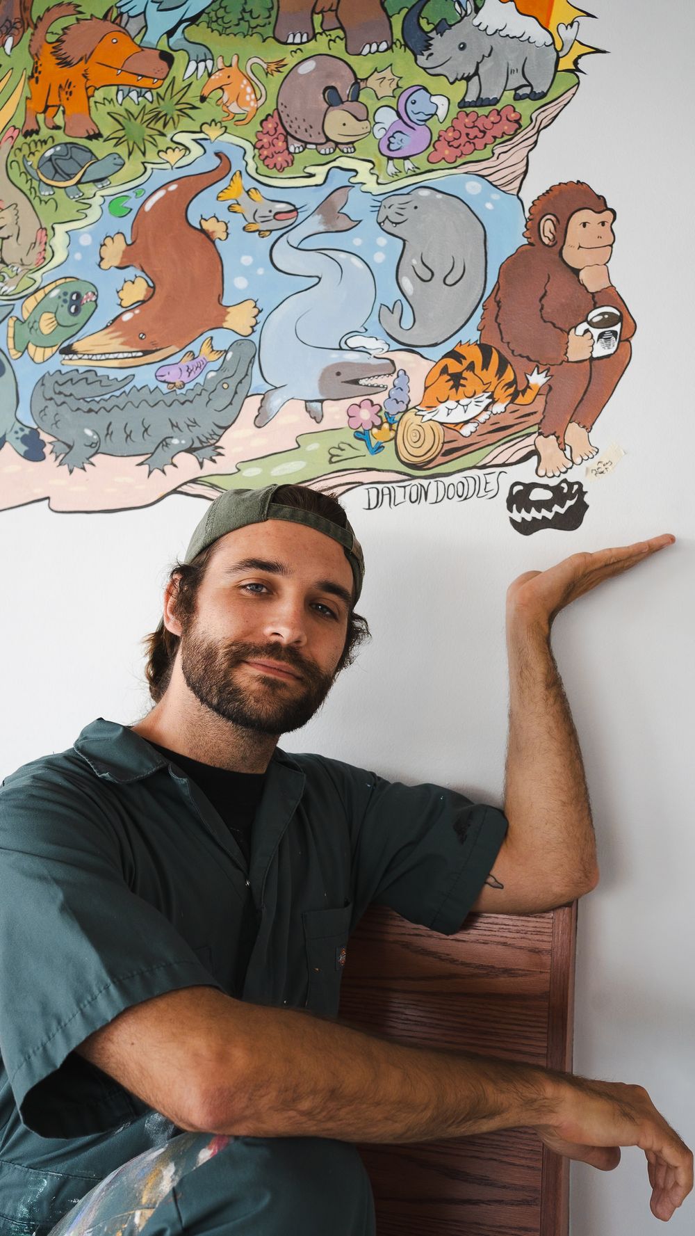 Artist gestures at colorful animal mural artwork featuring dinosaurs, elephants and monkeys on white wall.