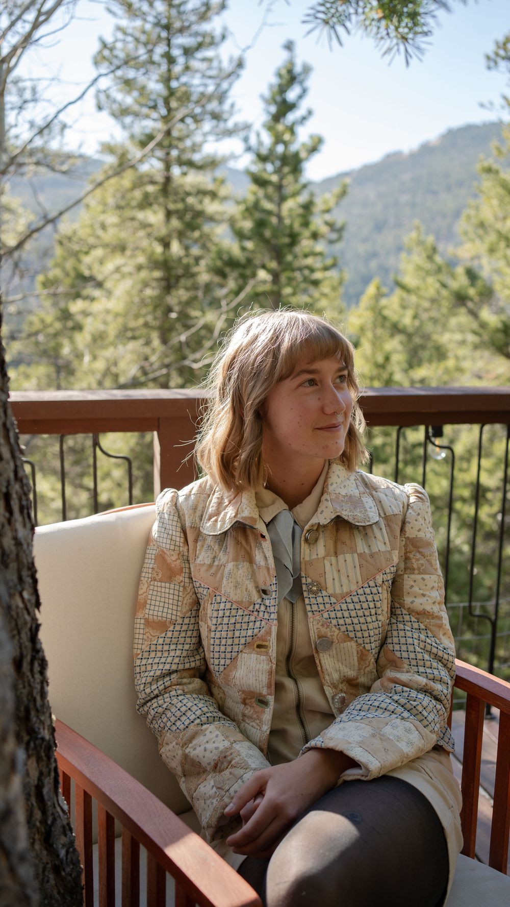 Person in patterned jacket sitting on outdoor deck with mountain views and pine trees.