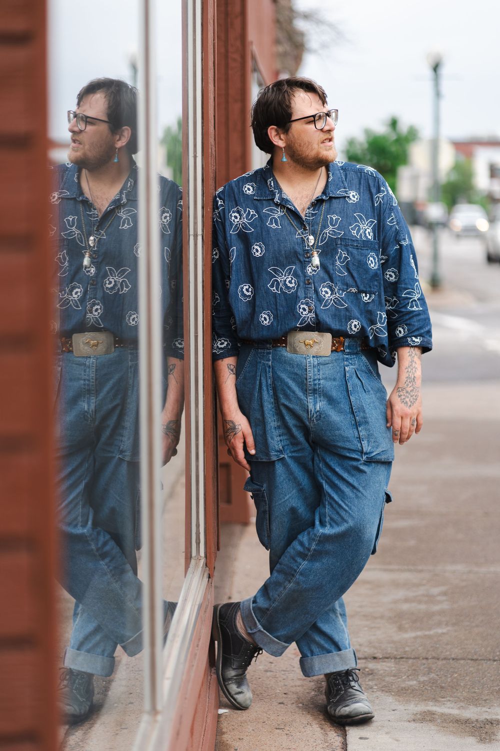 Person in blue patterned shirt and jeans leaning against reflective storefront window.