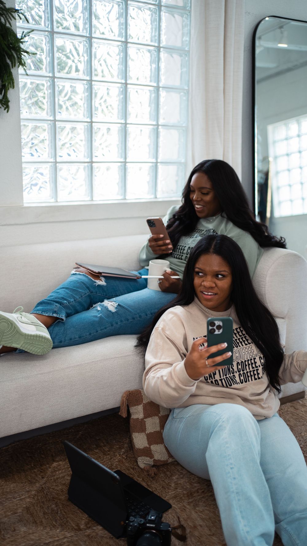 Two friends relaxing on white couch while checking their phones.